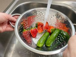 Strawberries and cucumber in a colander being rinsed with water in the sink 